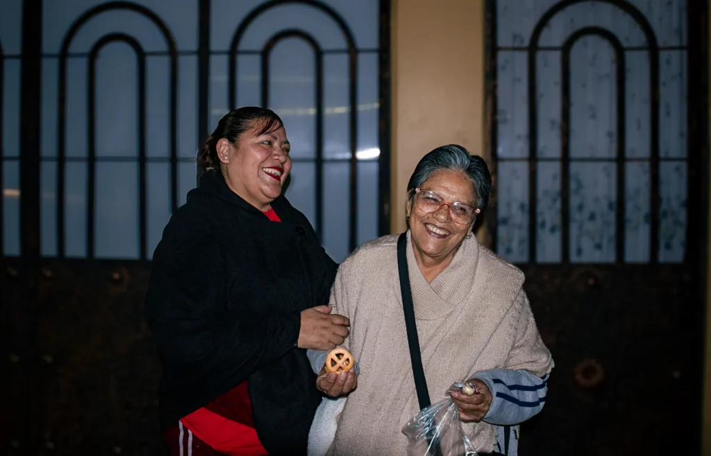 Dos mujeres sonríen y se ríen frente a una reja negra de noche; una de ellas sostiene una galleta o pan redondo
