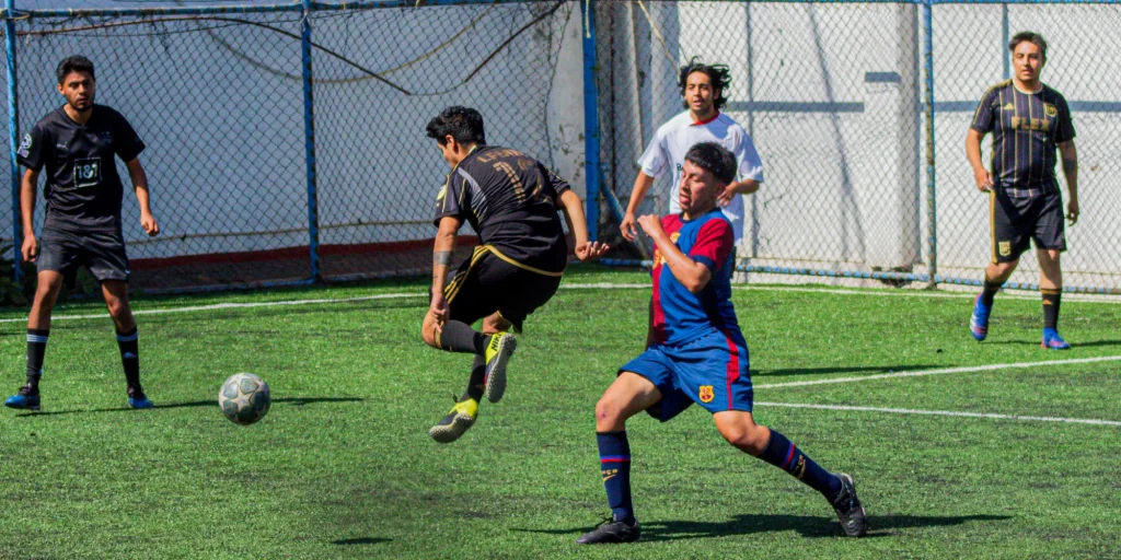 Jugadores disputan el balón en una cancha de pasto sintético: uno de negro salta a patear y otro de azul y rojo lo marca