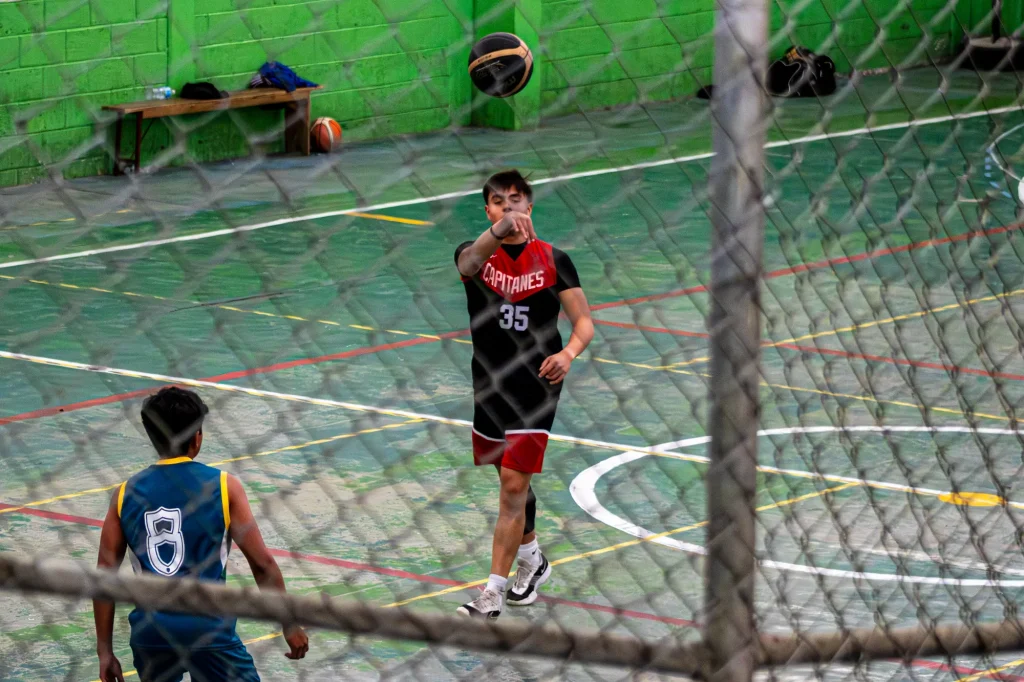 Jugador de basquetbol con camiseta roja y negra pasa el balón en una cancha vista a través de una malla