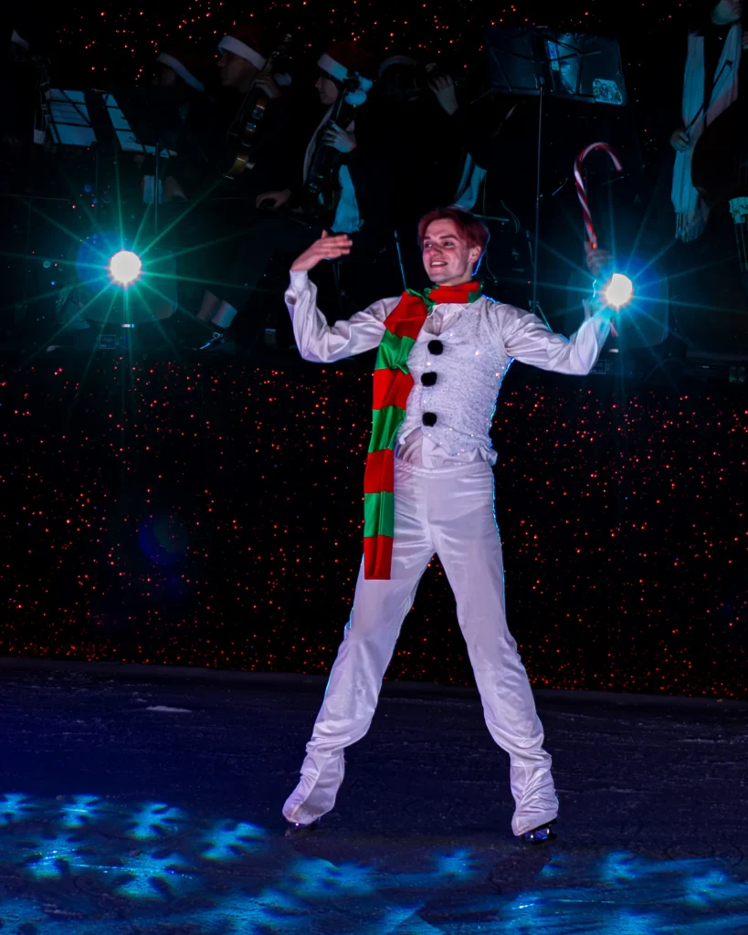 Artista con traje blanco de muñeco de nieve y bufanda roja y verde, con un brazo en alto en un escenario oscuro con luces