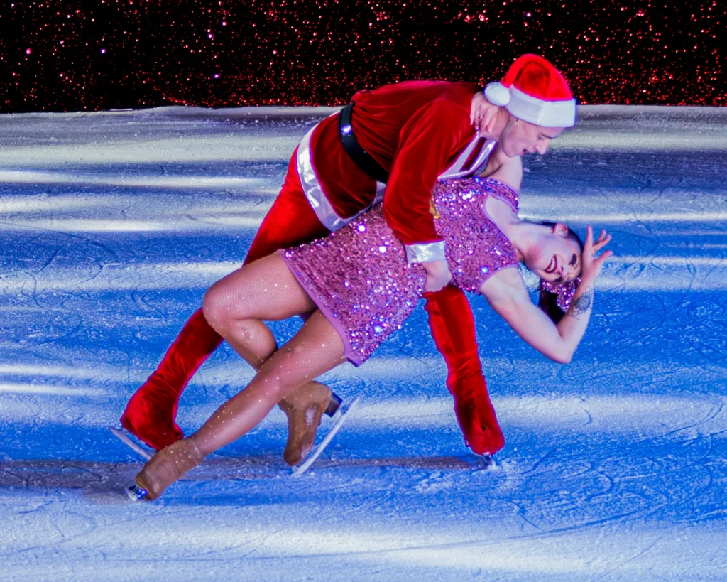 Pareja de patinadores sobre hielo con atuendo navideño: él, con gorro de Santa Claus, inclina a ella, de vestido rosa