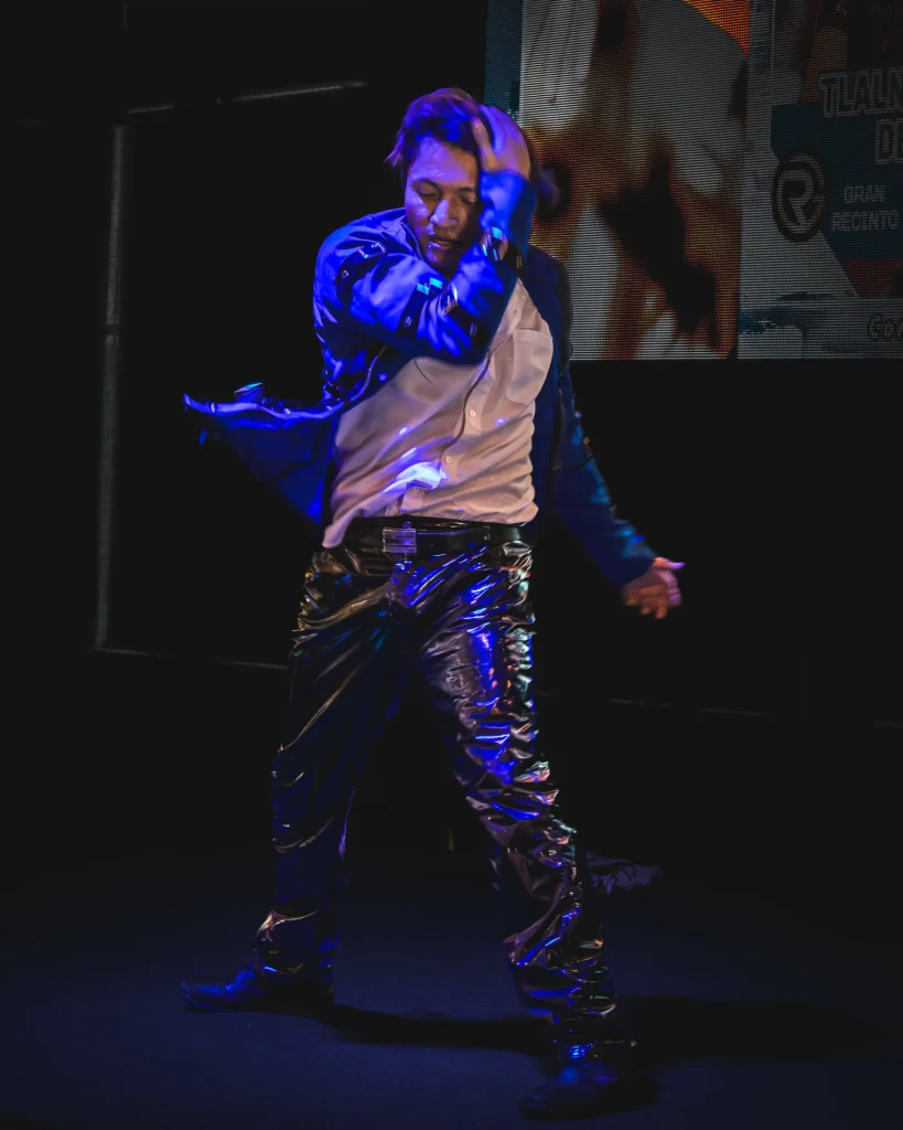 Hombre bailando en un escenario oscuro con luz azul, con chamarra azul, camisa blanca y pantalón negro brillante