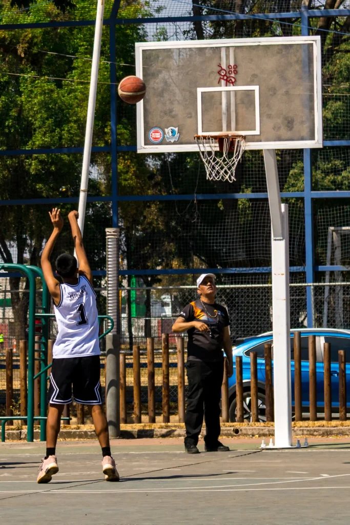 Jugador de basquetbol lanza un tiro hacia el aro en una cancha al aire libre, mientras una persona con gorra blanca observa