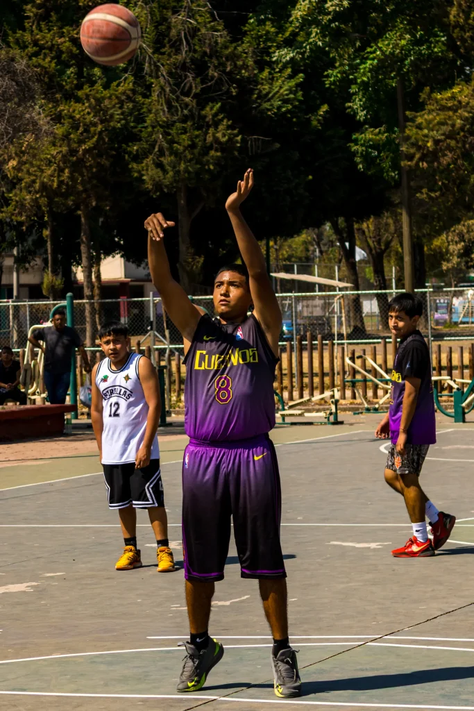 Jugador de uniforme morado con los brazos en alto justo después de lanzar un tiro; el balón sube mientras otros observan