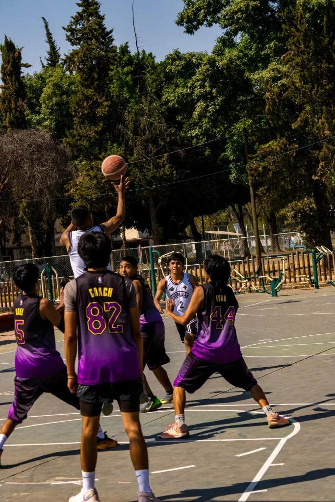 Jugadores de morado en primer plano mientras uno de blanco lanza un tiro al fondo, en una cancha rodeada de árboles