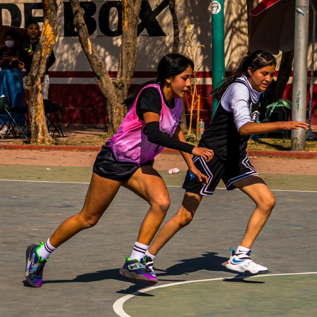 Dos jugadoras corren por la cancha: una con chaleco rosa y manga negra, otra de uniforme negro, ambas concentradas
