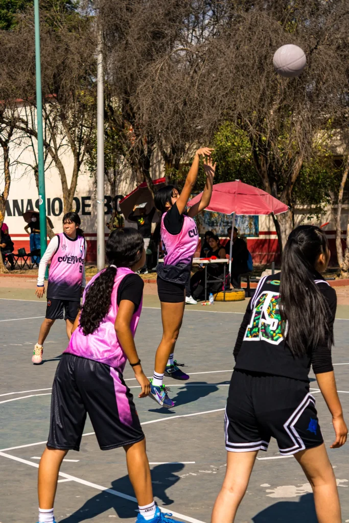 Jugadora con chaleco rosa lanza un tiro con el balón en el aire, mientras otras compañeras observan bajo los árboles