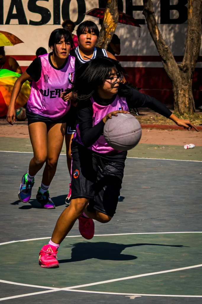 Jugadora con lentes y manga larga negra avanza botando el balón, seguida por dos compañeras con chalecos rosas