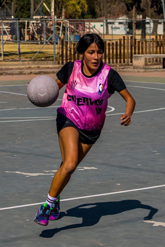 Jugadora con chaleco rosa bota el balón en carrera por una cancha al aire libre, con la mirada al frente