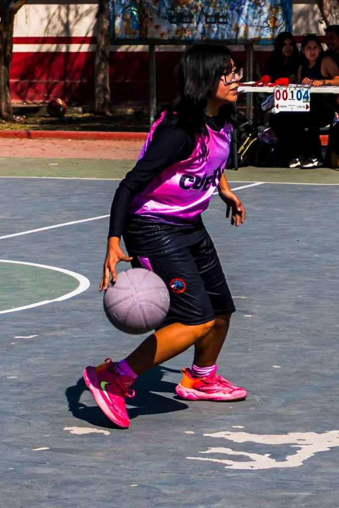 Jugadora con lentes y chaleco rosa bota el balón en la cancha; al fondo hay personas sentadas junto a una mesa