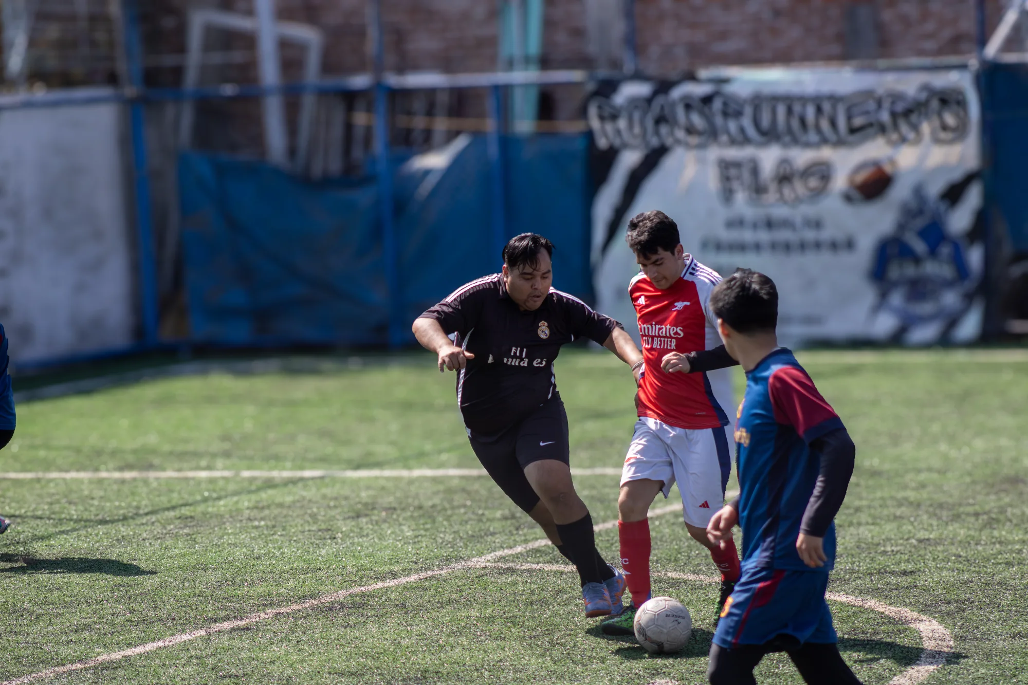 Tres jugadores de futbol disputan el balón en una cancha de pasto sintético, con una lona y una malla al fondo